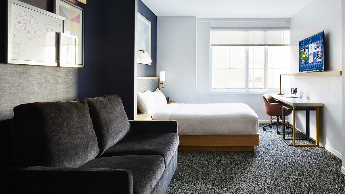 guest room at Club Quarters Hotel White House with carpeted floor, sofa with black uphostery and bed with white sheets and flat sceen tv on wall in Washington DC, USA